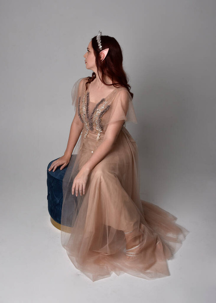 Volledig portret van een roodharig meisje met een romige fantasiejurk en kristallen kroon, als een sprookjesgodin kostuum. zittende houding met elegante gebaren handen, geïsoleerd op lichte studio achtergrond. - Foto, afbeelding