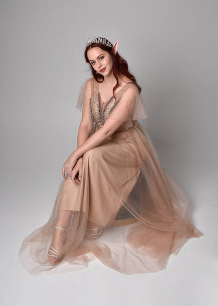 Full length  portrait of red haired  girl wearing a creamy fantasy gown and crystal crown, like a fairy goddess costume.  sitting pose with elegant gestural hands, isolated on light studio background. - Foto, Imagem