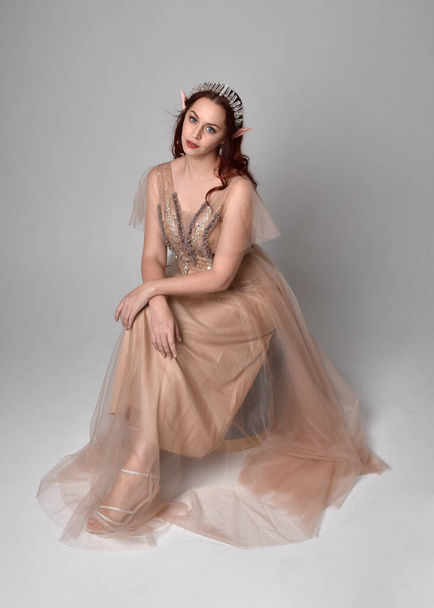 Full length  portrait of red haired  girl wearing a creamy fantasy gown and crystal crown, like a fairy goddess costume.  sitting pose with elegant gestural hands, isolated on light studio background. - Fotó, kép