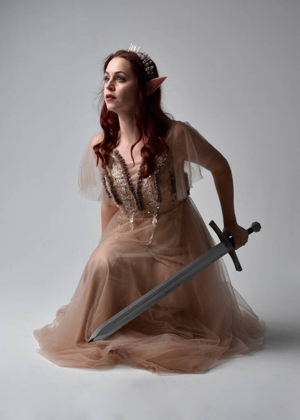 Полноразмерный портрет рыжеволосой девушки в кремовом фантазийном платье и хрустальной короне, как костюм богини-феи. сидя в позе с оружием, изолированным на светлом фоне студии. - Фото, изображение