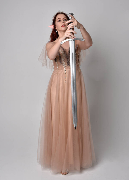 妖精の女神の衣装のように、クリーミーなファンタジーのガウンとクリスタルクラウンを身に着けている赤い髪の少女の完全な長さの肖像画。ライトスタジオの背景に隔離された剣の武器を保持立ちポーズ. - 写真・画像