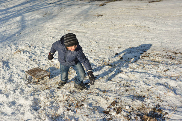 A fiú felmászik a hócsúszdán, és maga után húzza a szánkót. Téli hó játékok. - Fotó, kép