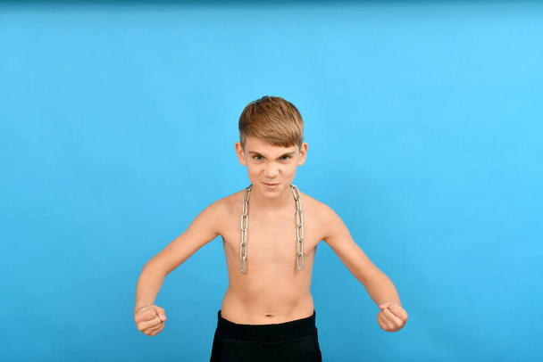Ein kleiner, dünner Junge mit einer Kette um den Hals wähnt sich als Bodybuilder. - Foto, Bild