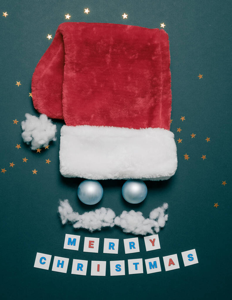 Рождественская открытка с красной кепкой Санты, голубыми шарами вместо глаз и усами из хлопка. Плоская композиция. Забавное лицо Санты - Фото, изображение
