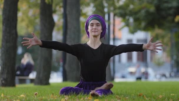 Ruhige, konzentrierte junge islamische Muslimin im Hijab sitzt im Park auf Gras im Freien und macht Yoga-Übungen Atemstrecktraining im Sommer auf Rasen Sport Flexibilität üben Pose - Filmmaterial, Video