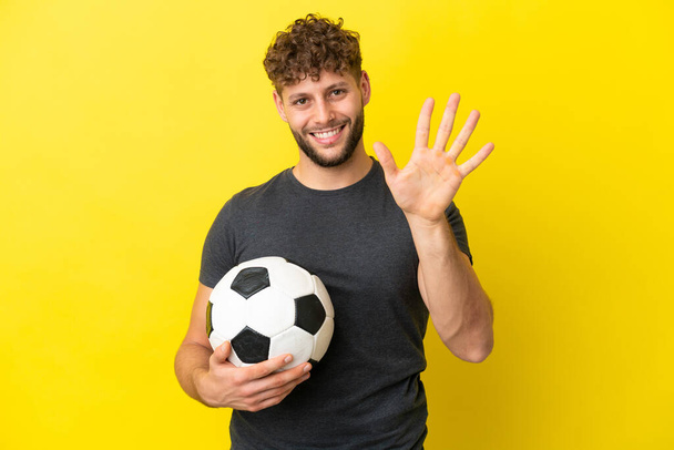 Όμορφος νεαρός ποδοσφαιριστής απομονωμένος στο κίτρινο φόντο μετρώντας πέντε με δάχτυλα. - Φωτογραφία, εικόνα