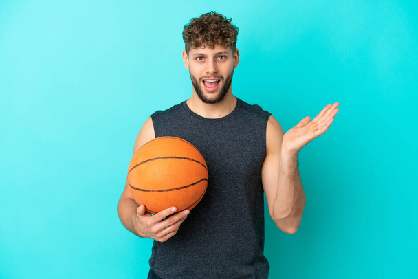 Όμορφος νεαρός που παίζει μπάσκετ απομονωμένος σε μπλε φόντο με σοκαρισμένη έκφραση προσώπου - Φωτογραφία, εικόνα