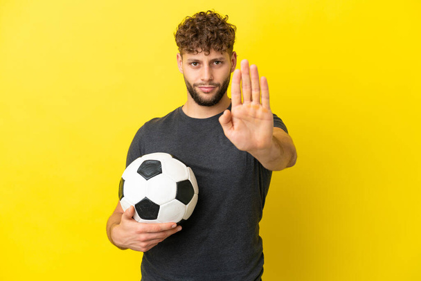 Όμορφος νεαρός ποδοσφαιριστής απομονωμένος σε κίτρινο φόντο κάνοντας χειρονομία stop - Φωτογραφία, εικόνα