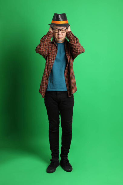 The senior Asian man dressing in Thanksgiving season color theme, wearing orange plaid shirt and black hat standing on the green background. - Photo, Image