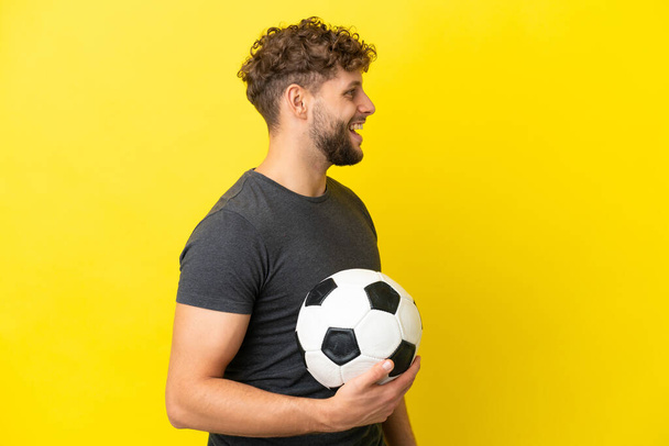 Knappe jonge football speler man geïsoleerd op gele achtergrond lachen in laterale positie - Foto, afbeelding