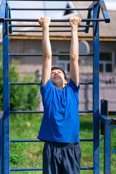 teenage boy exercising outdoors, sports ground in the yard, he pulls himself up on the horizontal bar, healthy lifestyle - Foto, afbeelding