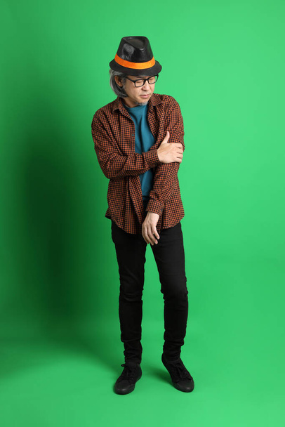 The senior Asian man dressing in Thanksgiving season color theme, wearing orange plaid shirt and black hat standing on the green background. - Foto, Bild