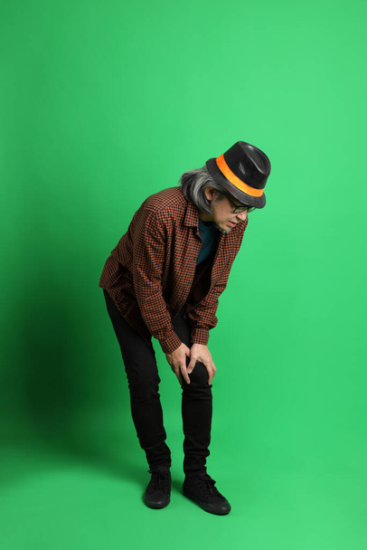 El hombre asiático mayor vestirse en el tema de color de la temporada de Acción de Gracias, con camisa a cuadros naranja y sombrero negro de pie sobre el fondo verde. - Foto, imagen