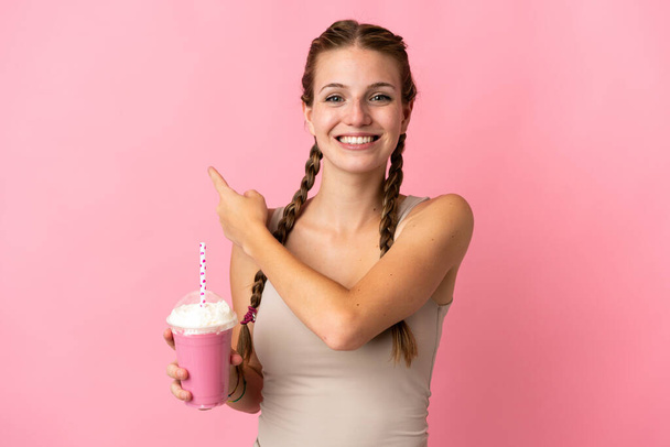 Jeune femme avec milkshake fraise isolé sur fond rose pointant vers le côté pour présenter un produit - Photo, image