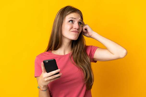 Young woman using mobile phone isolated on yellow background having doubts and with confuse face expression - Фото, зображення