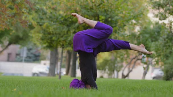 Молода сильна спортивна жінка в хіджабі і фіолетові штани зверху практикує на відкритому повітрі, тренер дівчини робить підтримувану позу голови, йога салама Сершасана Асан в парку влітку стоячи на голові на траві
 - Кадри, відео