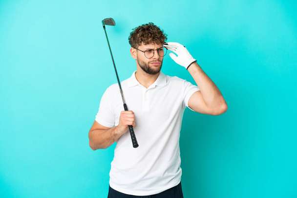 Mavi arka planda izole edilmiş yakışıklı beyaz bir adam golf oynuyor ve kafası karışmış bir yüz ifadesi var. - Fotoğraf, Görsel