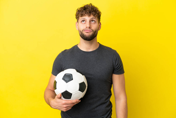 Όμορφος νεαρός ποδοσφαιριστής απομονωμένος σε κίτρινο φόντο και κοιτάζει ψηλά. - Φωτογραφία, εικόνα