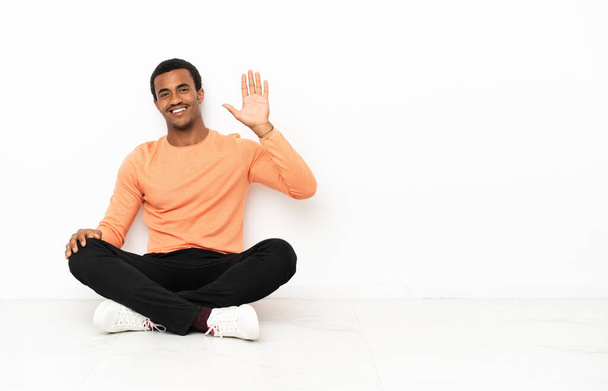 Hombre afroamericano sentado en el suelo sobre el fondo aislado del copyspace saludando con la mano con expresión feliz - Foto, Imagen
