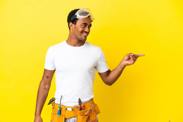 Afro-Amerikalı elektrikçi, izole edilmiş sarı duvarın üzerinde parmak göstererek bir ürün sunuyor. - Fotoğraf, Görsel