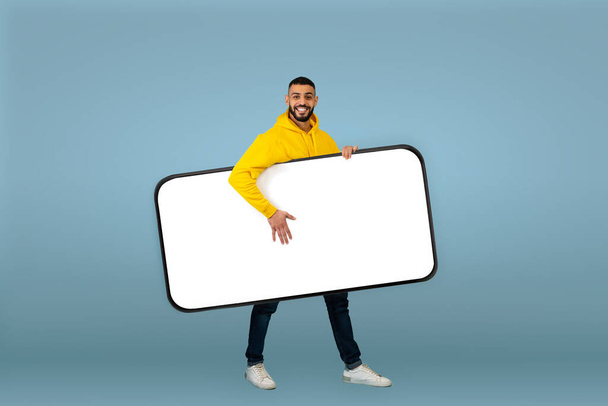Boldog arab férfi szállító nagy okostelefon üres fehér képernyő, séta a kütyü alatt kar, kék háttér - Fotó, kép