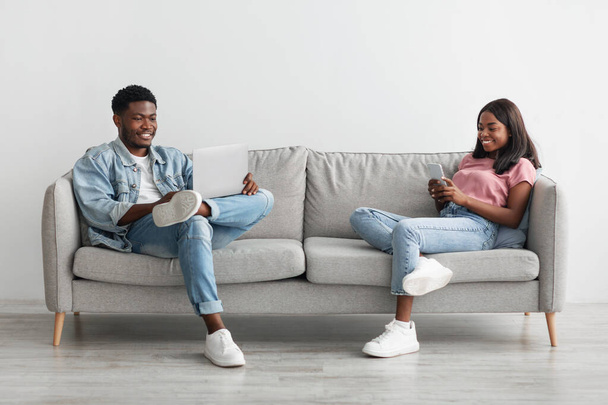 Happy black couple sitting on couch, using cellphone and pc - Photo, Image