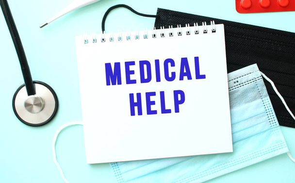 De blauwe tekst MEDICAL HELP is geschreven op een wit vel notitieblok dat op een blauwe achtergrond ligt naast een thermometer en medische gezichtsmaskers. Medisch concept - Foto, afbeelding