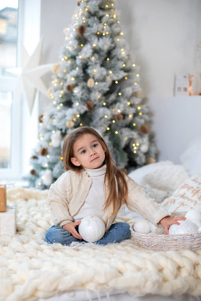 Happy little smiling girl with gift on Christmas Eve sit on bed. Child opens New year gift near christmas tree with glowing lights garland. Kid play in bed with xmas balls and toys in bedroom at home  - Photo, image