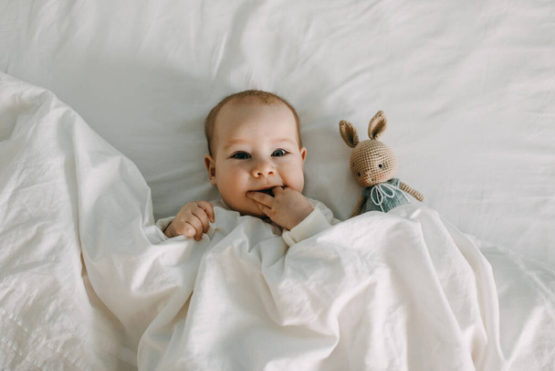 Onnellinen vauva makaa sängyssä valkoisen pehmeän peiton alla, hymyillen, imee sormia. - Valokuva, kuva