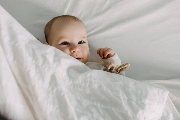 Primer plano de un bebé feliz acostado en la cama bajo una manta blanca y suave, sonriendo. - Foto, imagen