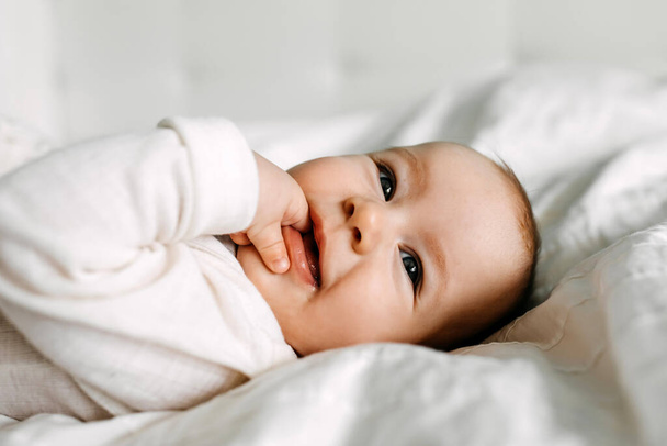 Sluiten van een gelukkige baby liggend in bed, glimlachend, zuigen vingers. - Foto, afbeelding