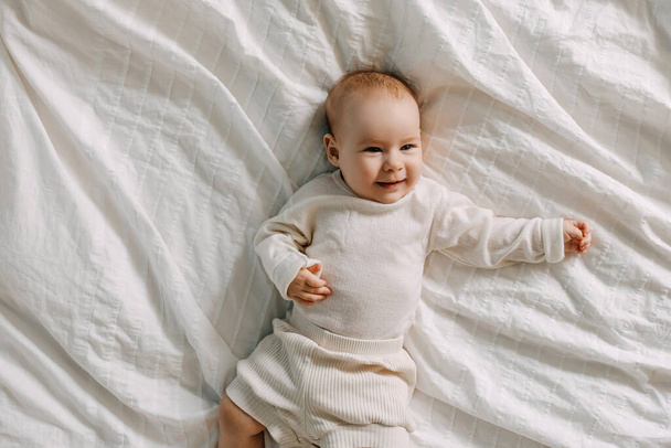 Five months old baby lying in bed on white bed sheet, smiling. - Фото, изображение
