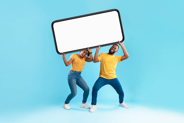 Emotional black couple holding big heavy smartphone with empty screen above their heads on blue background, mockup - Foto, imagen