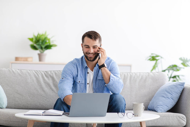 Vrolijke volwassen Europeaan met baard zitten op de bank werken thuis op afstand op laptop, bellen via de telefoon - Foto, afbeelding