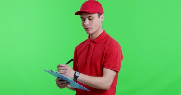 Young postman writing in check list, smiling at camera - Footage, Video