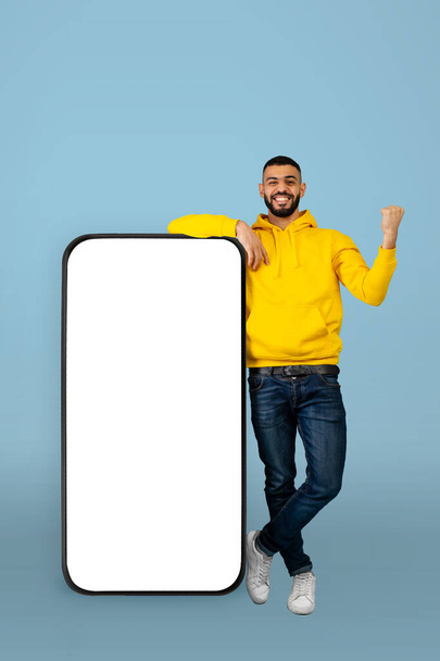 - Ναι. Ενθουσιασμένος άνδρας Άραβας ακουμπά στο smartphone με άδεια λευκή οθόνη και κουνώντας σφιχτή γροθιά, μπλε φόντο - Φωτογραφία, εικόνα