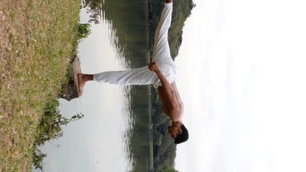 Бразильський юнак практикує пози йоги. Балансування на одній нозі в хмарний день на краю озера (вертикальне відео)). - Кадри, відео