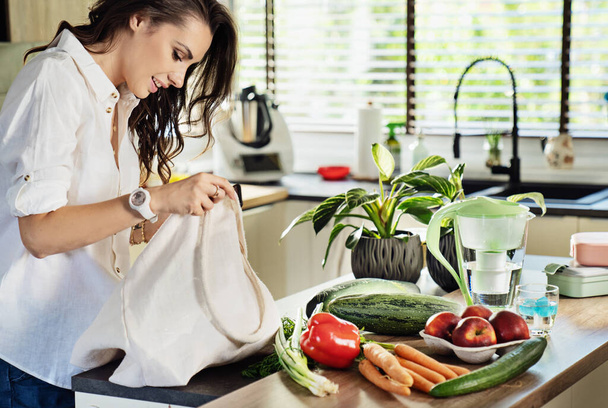 A beautiful young woman in a good mood sorting vegetables from a bag and putting them on the kitchen table - 写真・画像