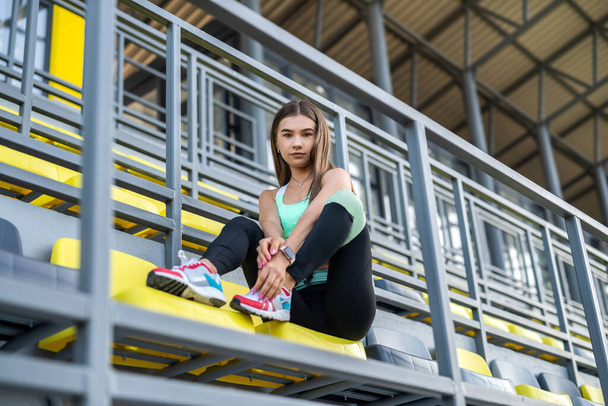 pretty slim woman in sport wear resting after hard workout near chairs on modern stadium. enjoy summer day - Photo, image