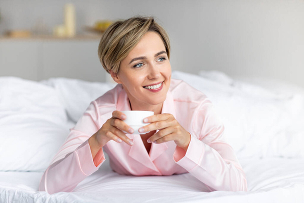 Happy woman looking away and thinking, drinking coffee in bed - 写真・画像