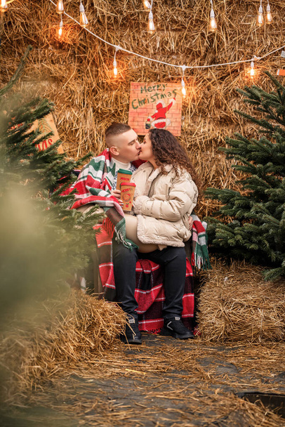 A beautiful young couple in white clothes are sitting near a Christmas tree in the garden. Happy man and woman, romance, Christmas celebration, fun, love. - Fotoğraf, Görsel