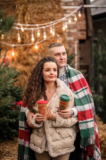 A beautiful young couple in white clothes are sitting near a Christmas tree in the garden. Happy man and woman, romance, Christmas celebration, fun, love. - Fotó, kép