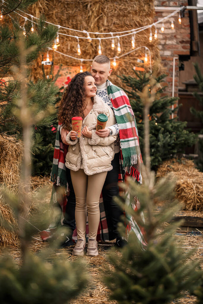 Ένα όμορφο νεαρό ζευγάρι με λευκά ρούχα κάθεται κοντά σε ένα χριστουγεννιάτικο δέντρο στον κήπο. Ευτυχισμένος άντρας και γυναίκα, ρομαντισμός, Χριστουγεννιάτικη γιορτή, διασκέδαση, αγάπη. - Φωτογραφία, εικόνα