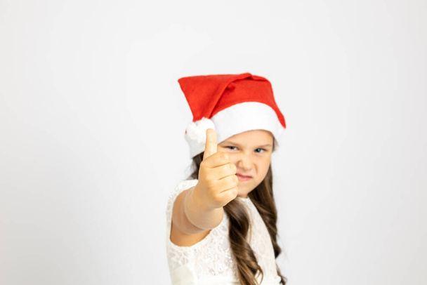 portrait of girl in white dress and wearing red Santa Claus hat gives thumbs up, isolated on white background with copy space - Фото, изображение