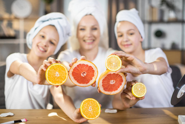 Woman with two daughters holding slices of citrus at table - Foto, afbeelding