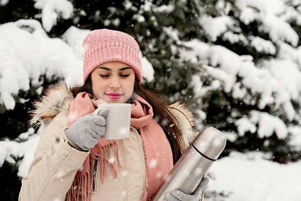 Vrolijk kerstfeest en gelukkig nieuwjaar. Mooie vrouw in warme winterkleding houden thermoskan en het drinken van warme thee of koffie buiten in besneeuwde dag - Foto, afbeelding