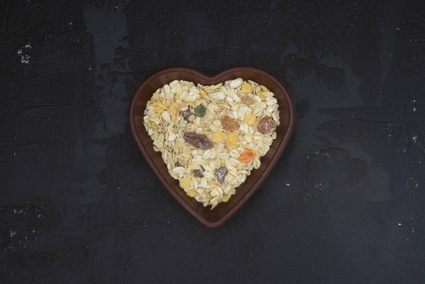 νιφάδες βρώμης και βρώμη σε πλάκα σχήματος καρδιάς σε μαύρο φόντο. Υγιές πρωινό. Βιολογικά και χορτοφαγικά τρόφιμα. Κομπόστα και δημητριακά. Επίπεδη lay, αντίγραφο χώρου για κείμενο - Φωτογραφία, εικόνα
