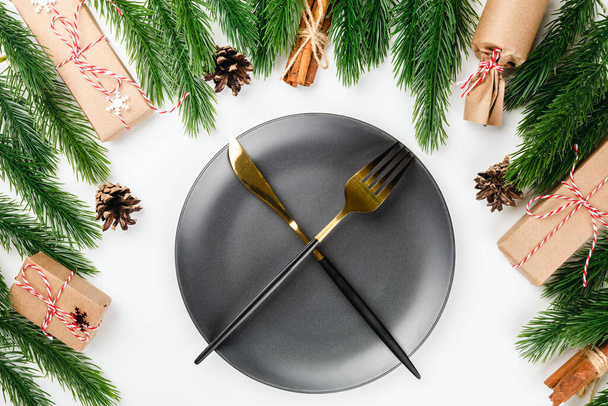 golden knife and fork crossed on black plate on Christmas dinner table with frame of fir branches, with cones and gifts - Foto, imagen