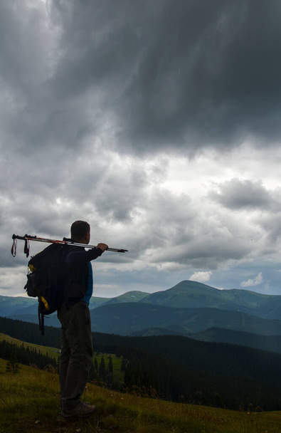 Caminante con mochila y bastones en la mano de pie y disfrutando de la vista del valle y las montañas durante el día nublado - Foto, imagen