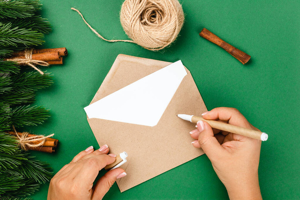 top view, process of writing letter to Santa Claus on craft envelope on green table with decorations of Christmas trees, cinnamon and jute rope - Photo, image